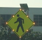 This photo shows a Pedestrian (W11-2) warning sign with 8 yellow illuminated LED lights around the border of the sign, with one LED light at each of the four corners of the sign and one LED light at each of the mid-points of the four sides of the sign.