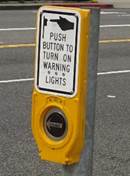 shows a pedestal-mounted pedestrian push button detector above which is mounted a PUSH BUTTON TO TURN ON WARNING LIGHTS (W10-25) sign.
