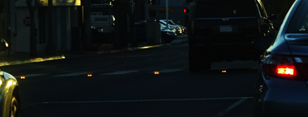 This photo shows a crosswalk that has been equipped with a series of yellow lights mounted on the pavement facing motorists.  The photo is dark because it was taken at night, but a set of four yellow lights facing the camera are illuminated and visible.
